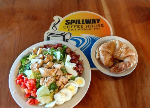 A plate of salad with chicken, tomatoes, and eggs, alongside a croissant, on a wooden table with a coffee house logo.