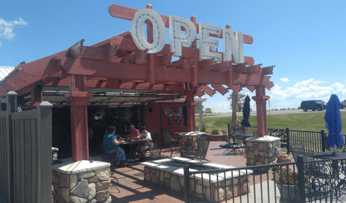 A rustic restaurant with an "OPEN" sign, outdoor seating, and patrons enjoying a meal under a clear blue sky.