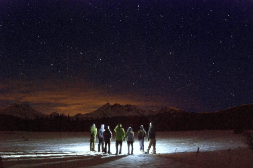 A group of people stands in a snowy landscape, illuminated by a flashlight, under a starry night sky.