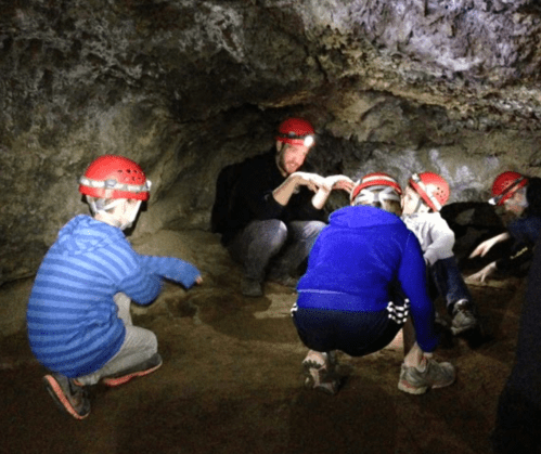 A group of children and an adult wearing helmets explore a cave, examining the ground in a dimly lit environment.