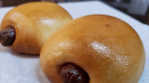 Two golden-brown, glossy buns with hot dogs peeking out, resting on a white surface.
