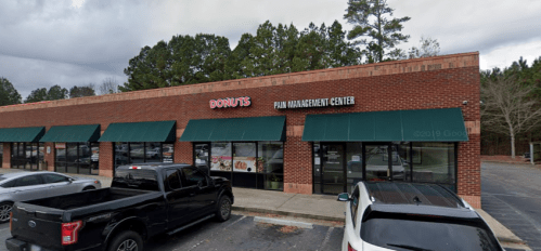A brick building with a donut shop and a pain management center, surrounded by trees and parked cars.