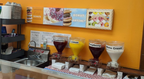 A colorful display of drink dispensers labeled Strawberry, Lemon, Raspberry, and White Cream, next to a donut sign.