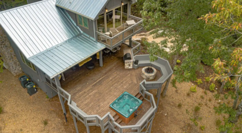 Aerial view of a modern house with a deck, hot tub, and fire pit, surrounded by trees and gravel pathways.