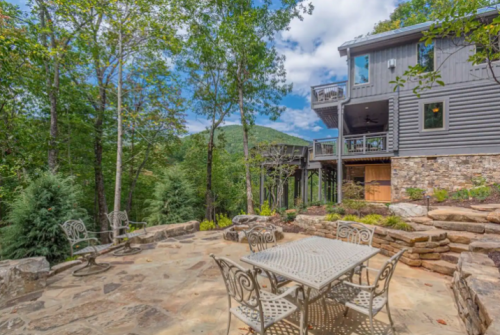 A stone patio with a metal table and chairs, surrounded by trees and a view of a mountain in the background.