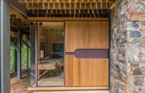 A modern wooden cabin with glass walls, nestled under a stone structure in a natural setting.