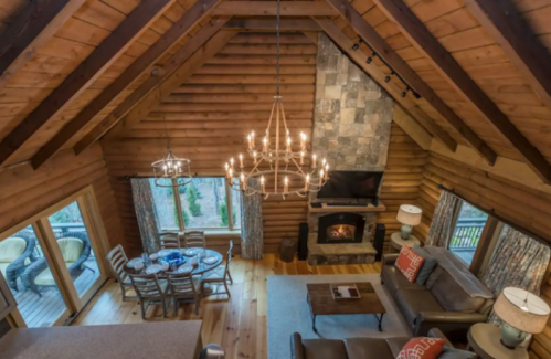 Cozy log cabin interior with a chandelier, stone fireplace, and rustic furniture, featuring large windows and a dining area.