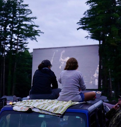 Two people sit on a car roof, watching a movie on an outdoor screen surrounded by trees at dusk.