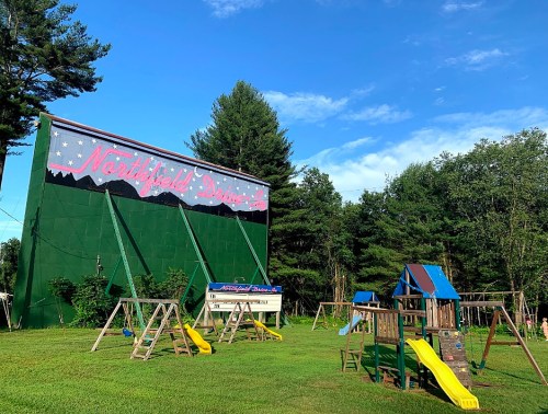 A green drive-in theater sign with "Northfield Drive-In" and a playground featuring swings and slides in the foreground.