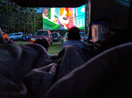 A cozy outdoor movie scene from inside a vehicle, with a colorful screen and people watching from under blankets.