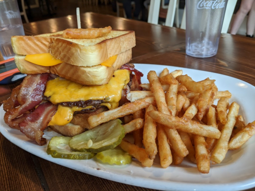 A towering sandwich with bacon, cheese, and toast, served with a side of crispy fries and pickles.