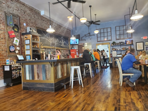 A cozy restaurant interior with a wooden bar, brick walls, and patrons dining at tables. Brightly lit and inviting atmosphere.