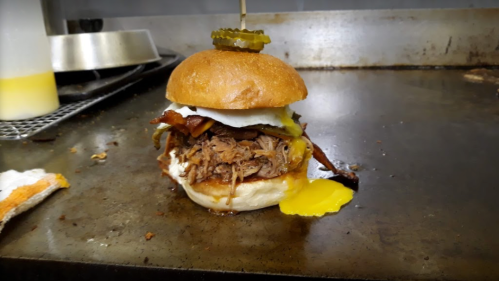 A delicious burger with pulled pork, bacon, a fried egg, pickles, and a golden bun, resting on a grill surface.