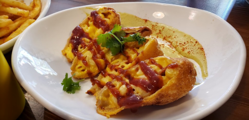 A plate of crispy mac and cheese bites drizzled with sauce and garnished with cilantro, served with a green dip.