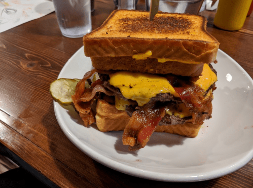 A stacked burger with two beef patties, melted cheese, bacon, and pickles, served between two slices of toasted bread.