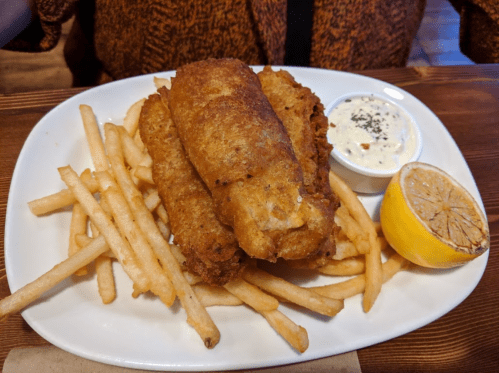 A plate of golden-brown fish fillets with fries, a lemon wedge, and a side of dipping sauce.