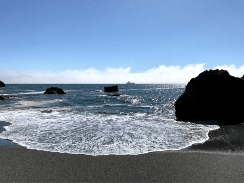 A serene beach scene with gentle waves lapping at dark rocks under a clear blue sky. A ship is visible in the distance.