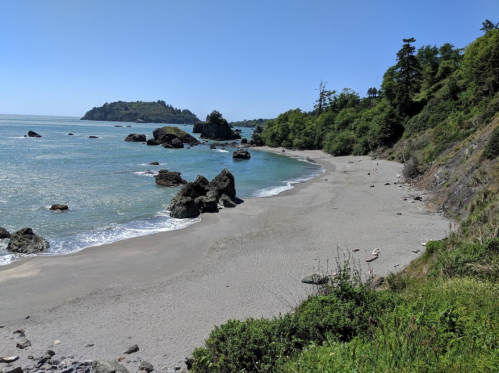 A serene beach scene with rocky outcrops, gentle waves, and lush greenery under a clear blue sky.