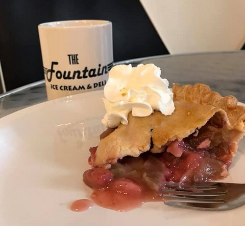 A slice of pie topped with whipped cream on a plate, next to a coffee mug that reads "The Fountain Ice Cream & Deli."