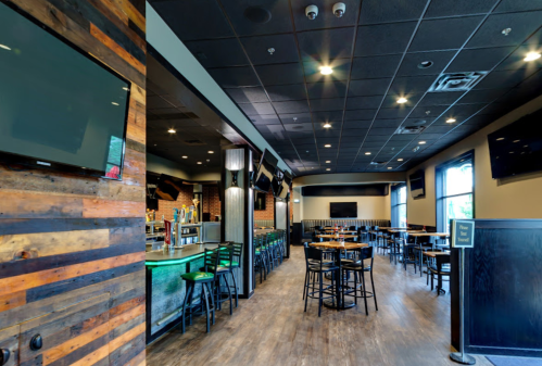 Interior of a restaurant with wooden walls, bar area, tables, and multiple TVs on the walls. Brightly lit and spacious.