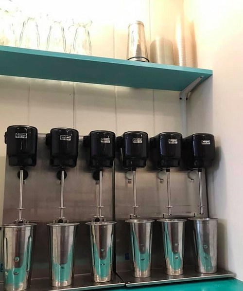 A row of black drink dispensers mounted on a wall above stainless steel cups on a turquoise shelf.