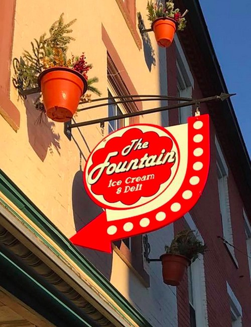 Sign for "The Fountain Ice Cream & Deli" with decorative pots and a vintage design, hanging from a building.