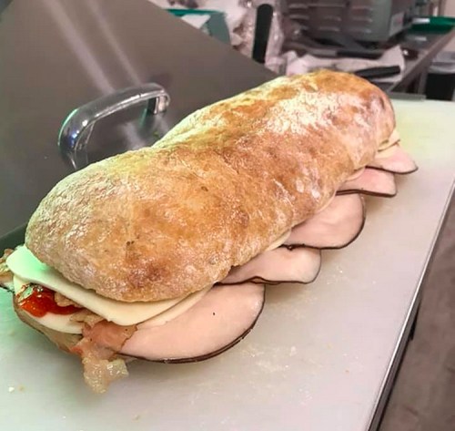 A large sandwich on a cutting board, filled with layers of meat, cheese, and sauce, in a rustic bread roll.