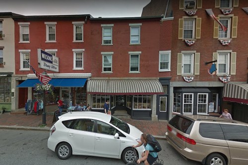 A street view of a quaint town with brick buildings, shops, and a parked car in front of a storefront.