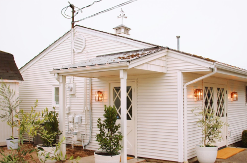 A charming white house with a porch, surrounded by potted plants, under a cloudy sky.