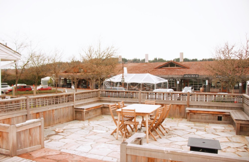 Outdoor seating area with wooden tables and chairs, surrounded by trees and buildings in a rustic setting.