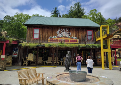 A rustic wooden building with a bear statue outside, surrounded by trees and outdoor seating. Visitors are nearby.
