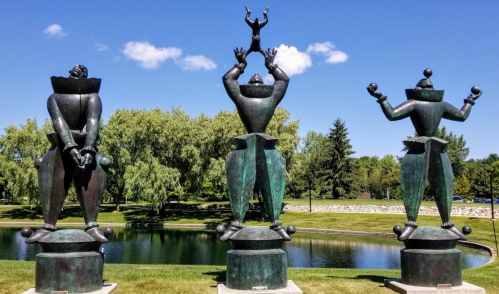 Three large bronze clown sculptures stand by a pond, each striking a different pose against a blue sky and green trees.