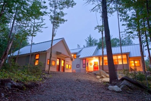 A modern house in a wooded area, illuminated at dusk, with large windows and a gravel path leading to the entrance.