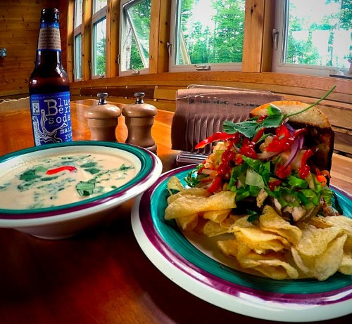 A plate with a sandwich topped with greens and salsa, a bowl of soup, chips, and a bottle of blueberry soda on a wooden table.