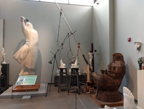 A gallery display featuring various sculptures, including a large figure, wooden art, and tools, in a well-lit space.
