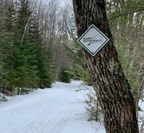 A snowy trail in a forest with a sign for Maine Huts & Trails attached to a tree.