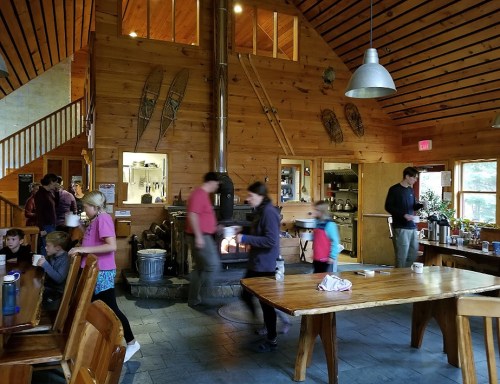 A cozy wooden lodge interior with people socializing, a fireplace, and a large dining table.