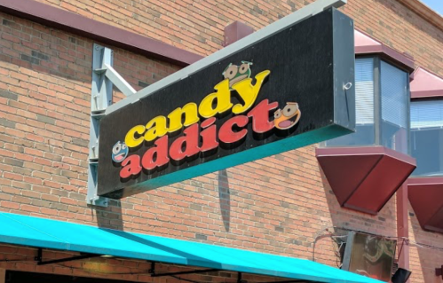 Sign for "Candy Addict" store with colorful lettering, mounted on a brick building with a teal awning.