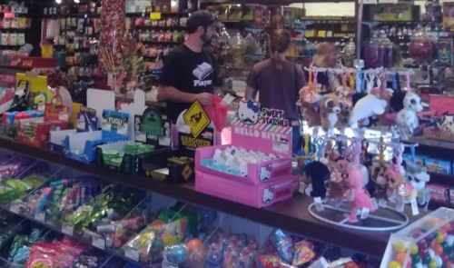 A colorful candy store display with various sweets, toys, and two customers browsing in the background.