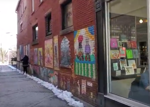 A colorful mural on a brick wall beside a bookstore, with snow along the sidewalk and a person walking by.