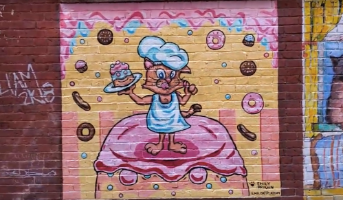 A colorful mural of a cat chef holding a cake, surrounded by donuts and sprinkles on a pink and yellow background.