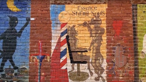 Mural depicting a barber and a customer, with vibrant colors and the text "Essence Styling Souls" on a brick wall.