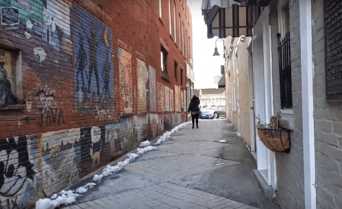 A person walks down a narrow alley lined with colorful murals and snow patches, with buildings on either side.