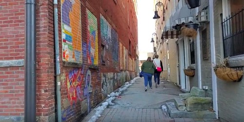 Two people walk down a narrow alleyway lined with colorful murals and brick walls, with sunlight streaming in.