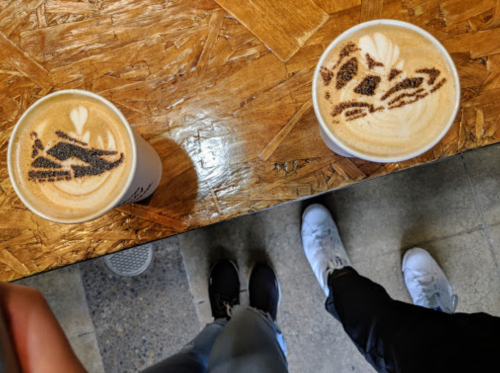 Two cups of latte art on a wooden table, with shoes visible below. One cup features a heart design.