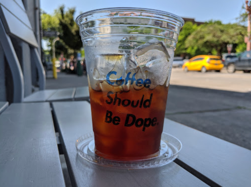 A clear cup of iced coffee with the text "Coffee Should Be Dope" on a table, with a street view in the background.