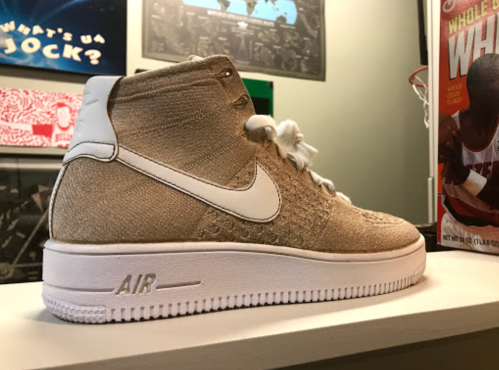 A gold Nike Air Force 1 sneaker displayed on a shelf, featuring a high-top design and white accents.