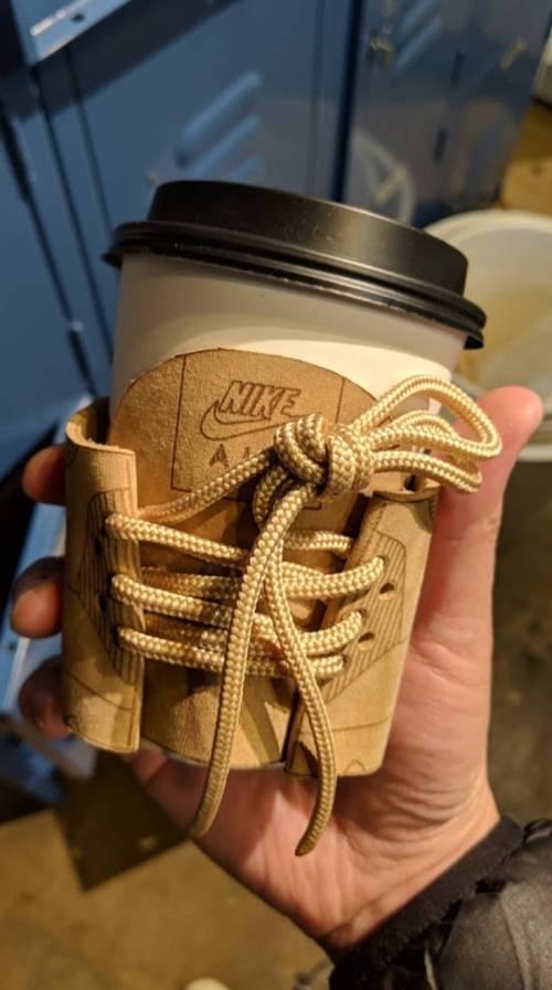A hand holding a coffee cup with a unique Nike-themed sleeve designed to look like shoelaces.