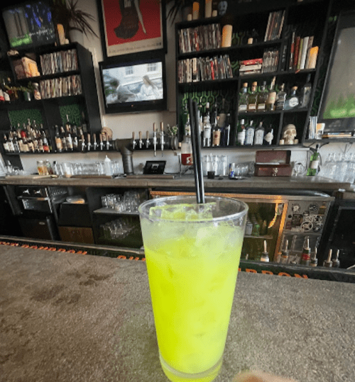 A vibrant green cocktail sits on a bar counter, with shelves of liquor and vintage decor in the background.