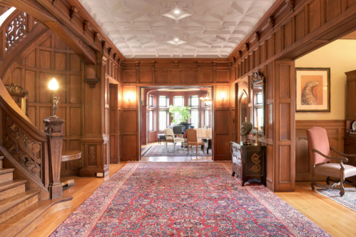 A grand wooden foyer with intricate details, a staircase, and a view into a bright, elegant dining area.